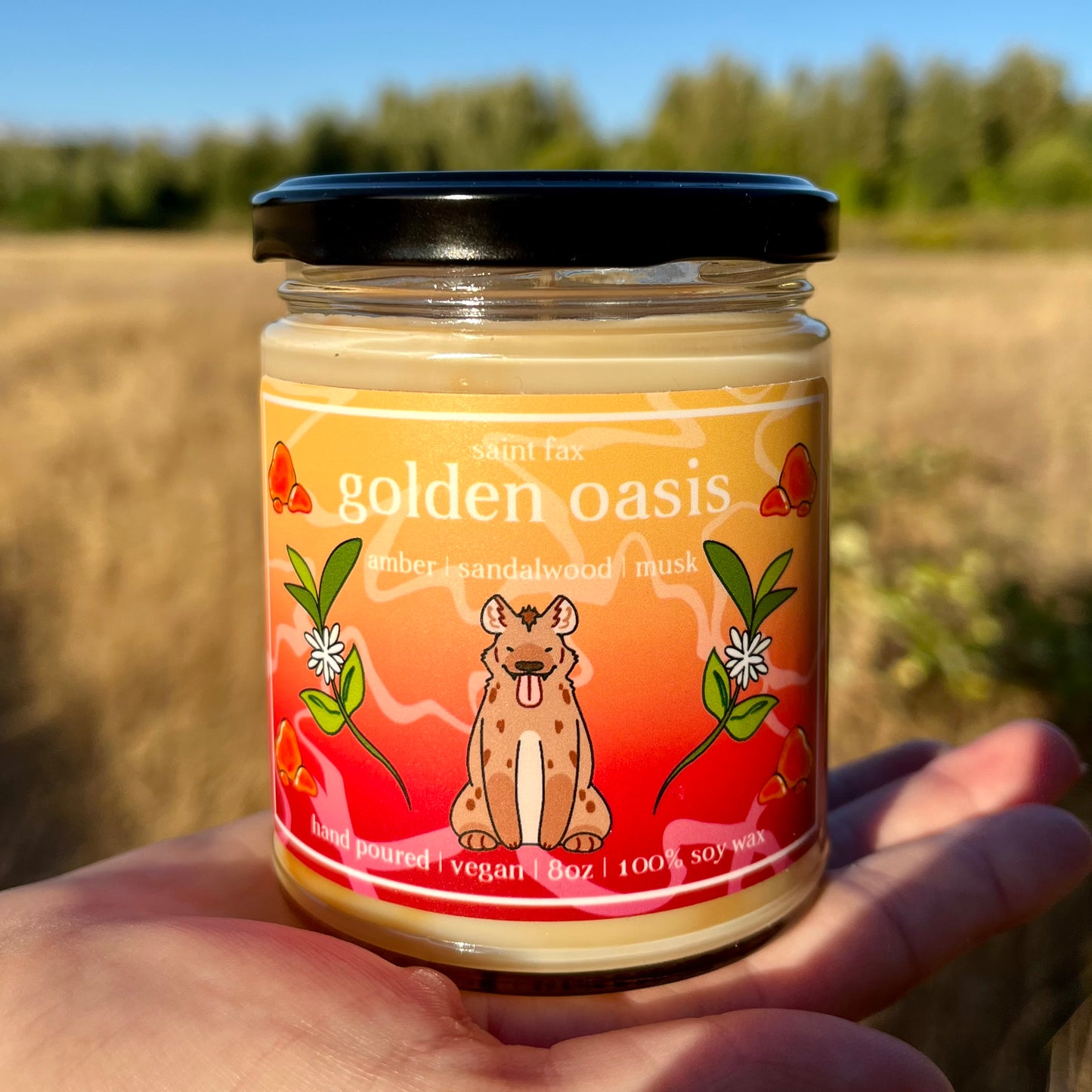 A jarred candle with a black lid and an orange/yellow gradient label with an illustration of a hyena surrounded by amber and sandalwood blossoms
The label reads:

saint fax
golden oasis
amber | sandalwood | musk
hand poured | vegan | 8oz | 100% soy wax