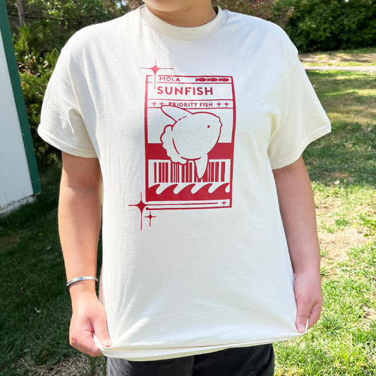 Model 1 wearing a light cream t-shirt with a red Sunfish graphic styled like a postal label while outdoors