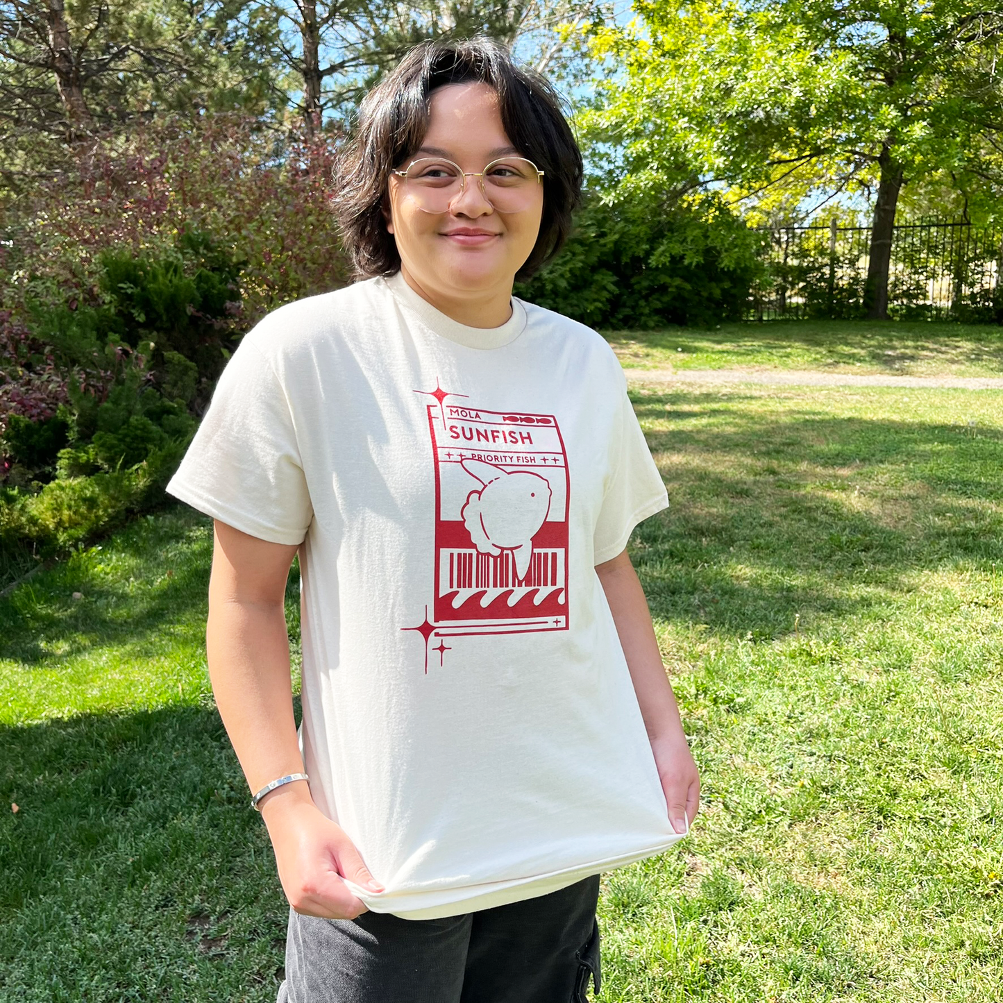 Model 1 wearing a light cream t-shirt with a red Sunfish graphic styled like a postal label while outdoors