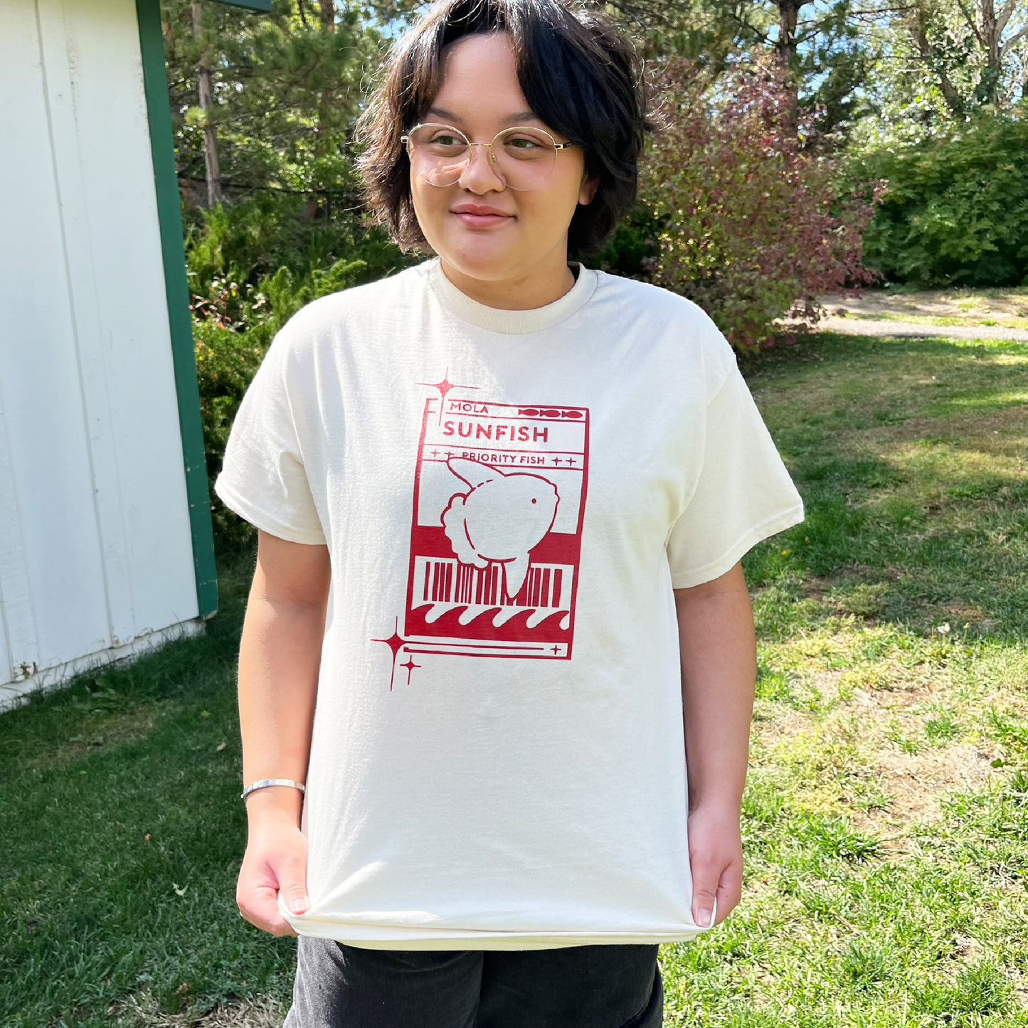 Model 1 wearing a light cream t-shirt with a red Sunfish graphic styled like a postal label while outdoors