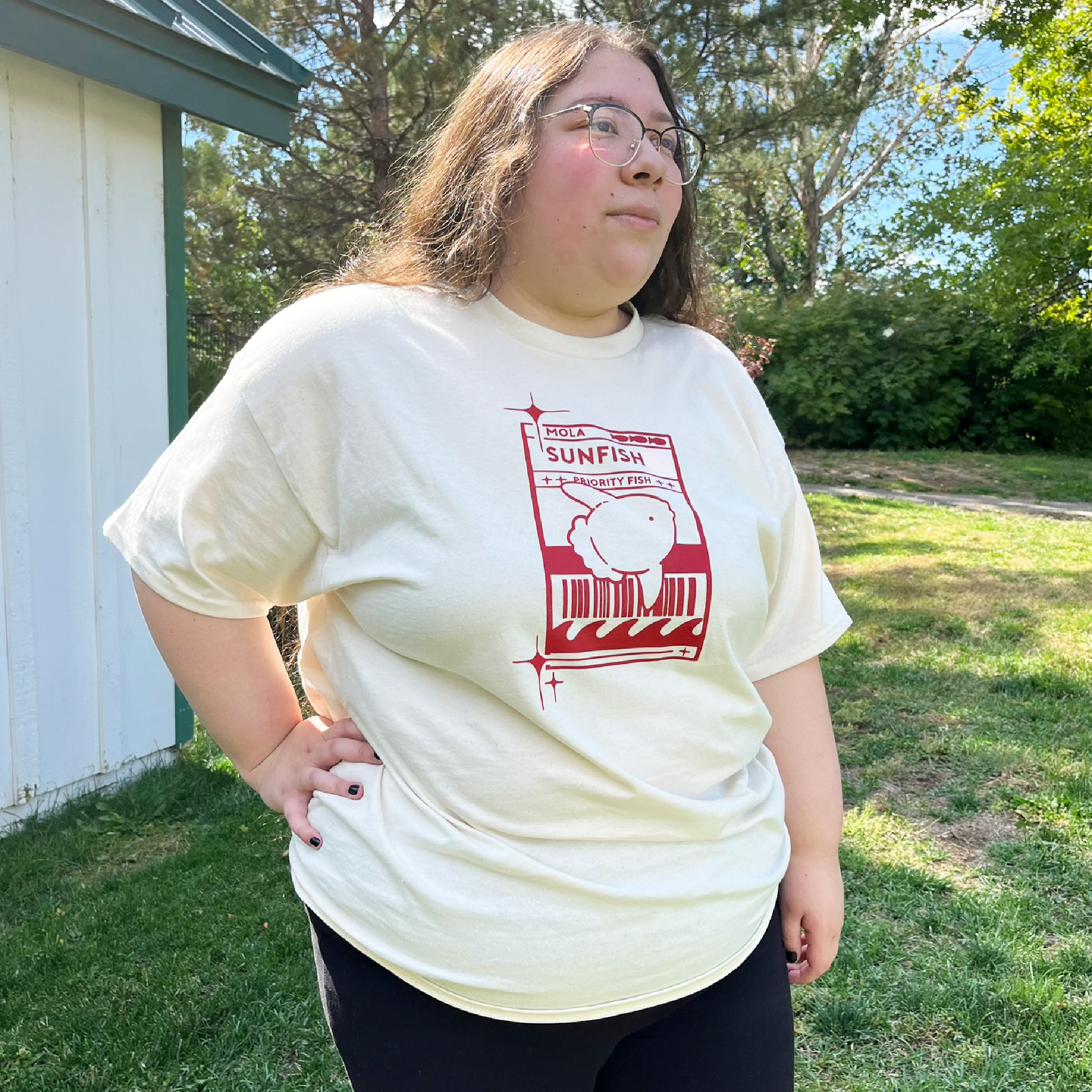 Model 2 wearing a light cream t-shirt with a red Sunfish graphic styled like a postal label while outdoors