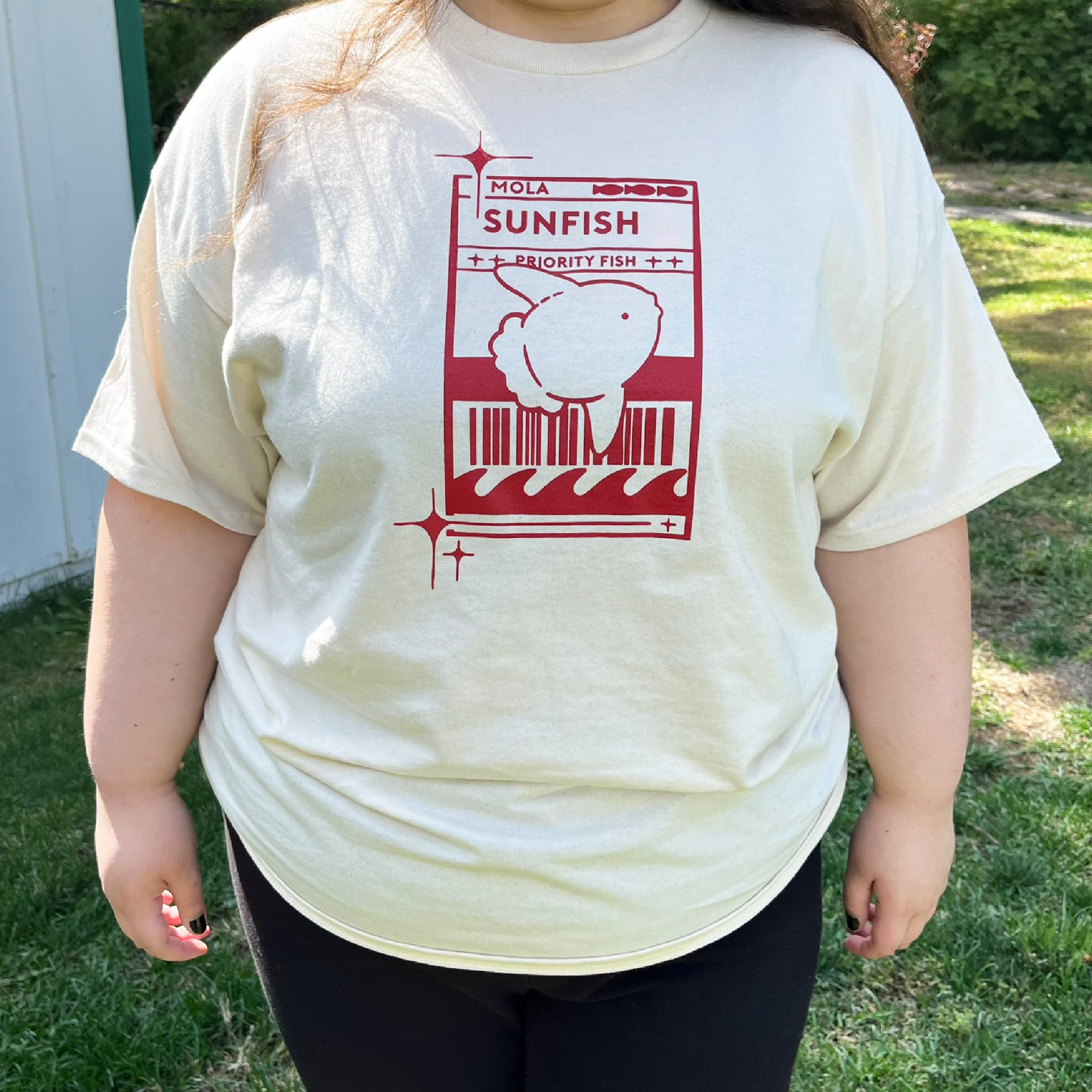 Model 2 wearing a light cream t-shirt with a red Sunfish graphic styled like a postal label while outdoors